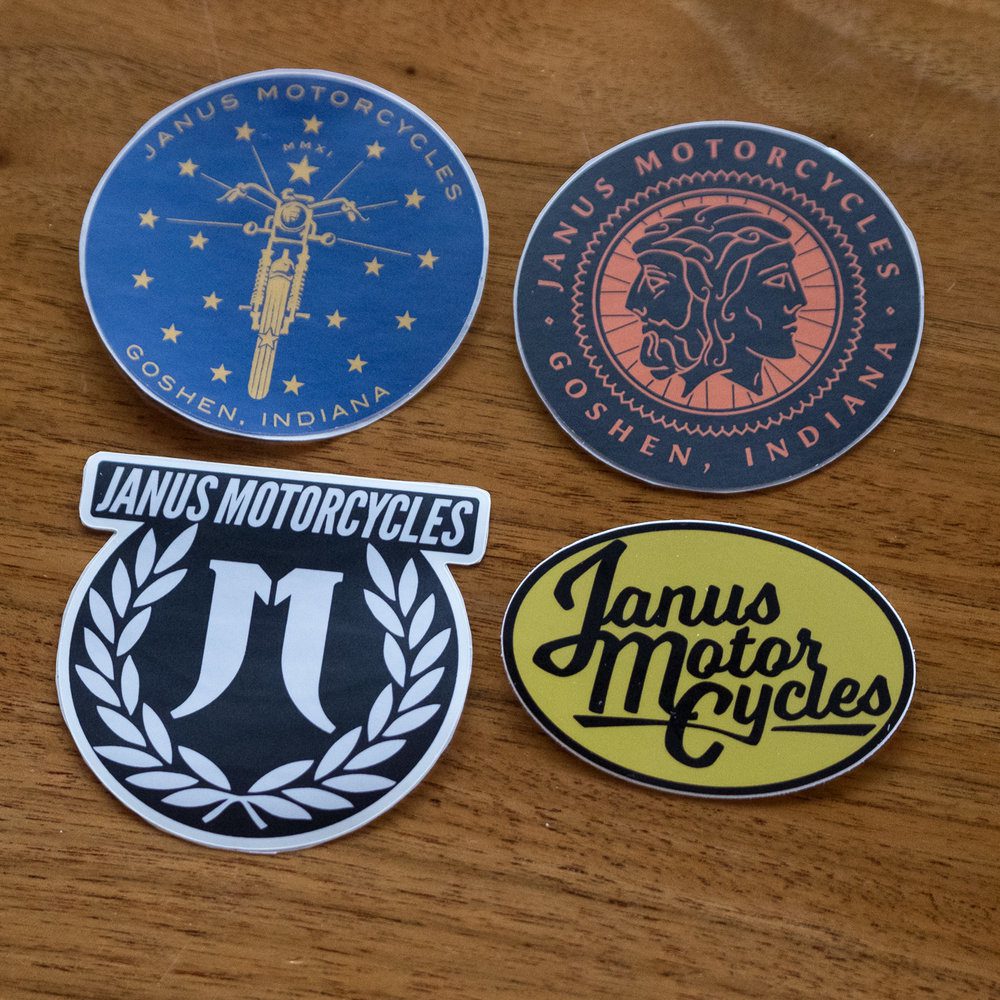 Alt: Collection of four Janus Motorcycles stickers on a wooden surface.
