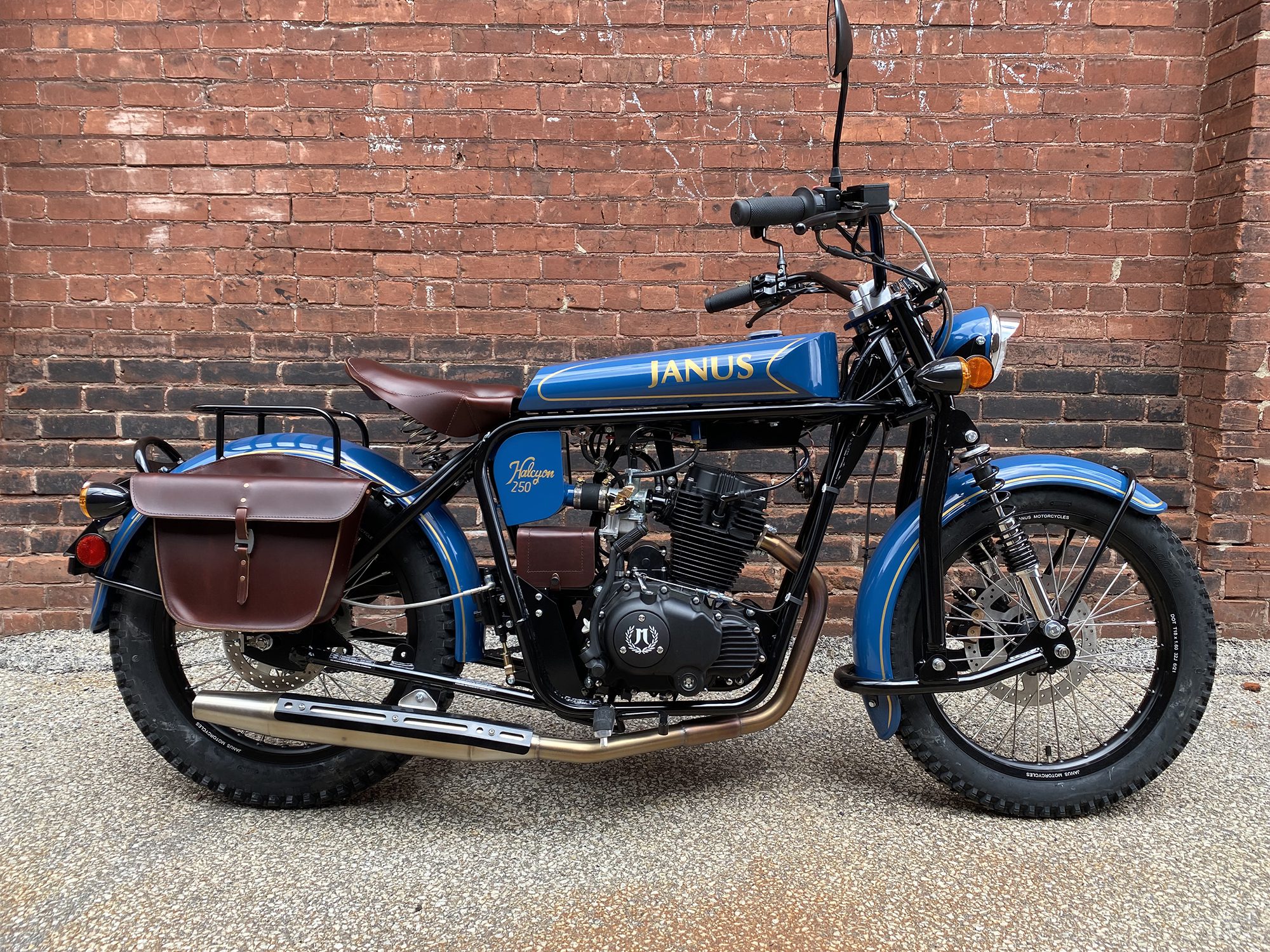 Blue Janus motorcycle with leather saddlebags parked against a brick wall.