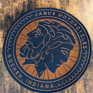 Round Janus Motorcycles branded coaster with a two-faced profile on a wooden table.